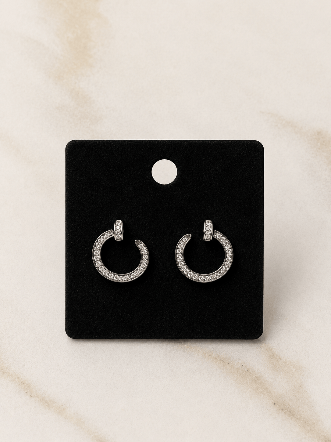 A pair of silver crescent-shaped hoop earrings paved with small clear stones, displayed on a black rectangular card against a light beige marble background.