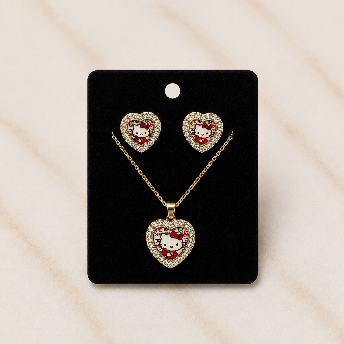A gold heart-shaped jewelry set featuring a pendant necklace and two matching stud earrings, displayed on a black card. The heart pendants and studs are decorated with clear rhinestones and feature a white Hello Kitty face with a red bow, set against a light beige marble background.