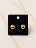 A pair of small gold ball stud earrings displayed on a black rectangular card against a light beige marble-like background.