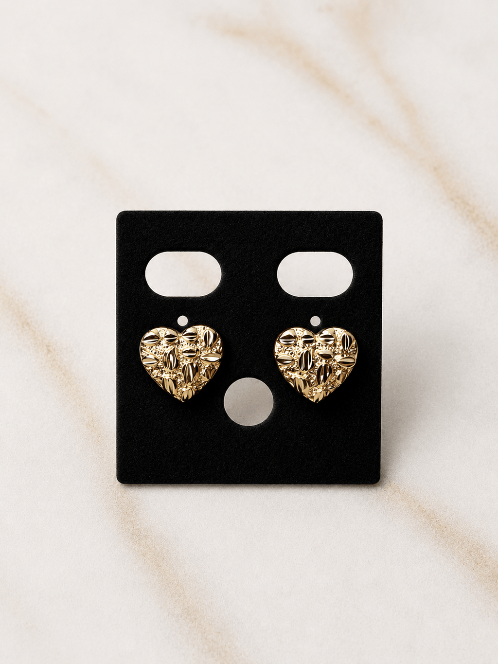 A pair of textured gold heart stud earrings displayed on a black rectangular jewelry card, set against a light beige marble background.