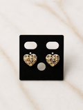 A pair of textured gold heart stud earrings displayed on a black rectangular jewelry card, set against a light beige marble background.