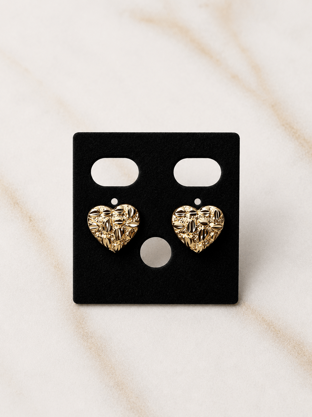 A pair of textured gold heart stud earrings displayed on a black rectangular jewelry card, set against a light beige marble background.