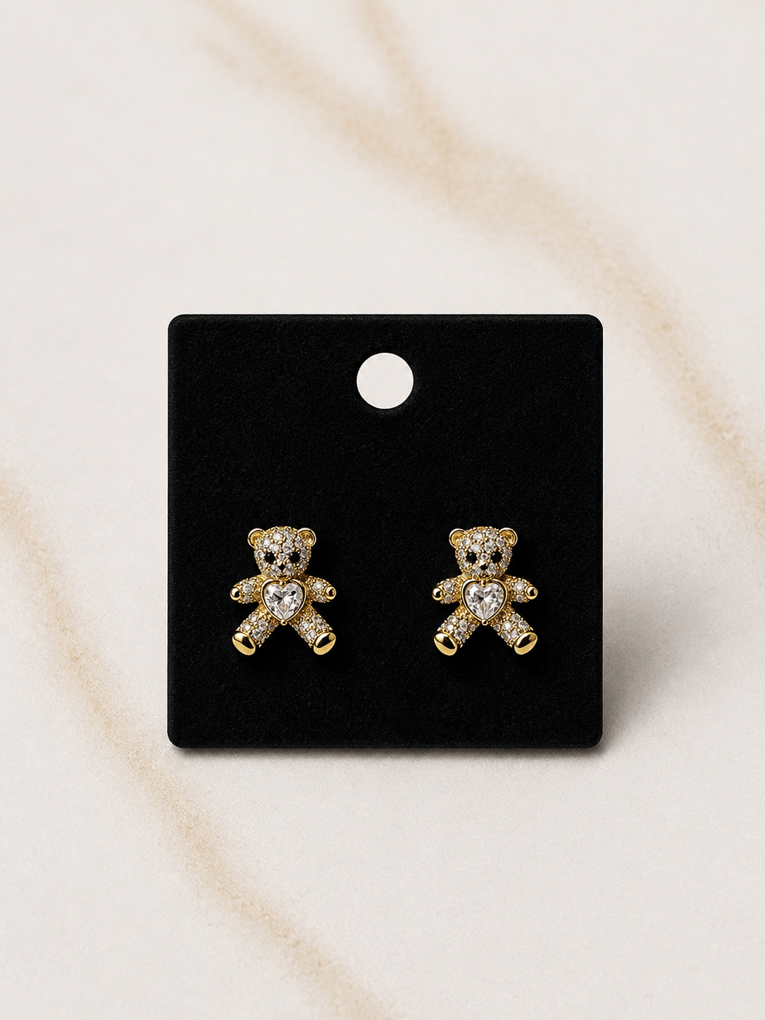 A pair of gold teddy bear stud earrings with clear crystal details, displayed on a black rectangular card against a light beige marble background.
