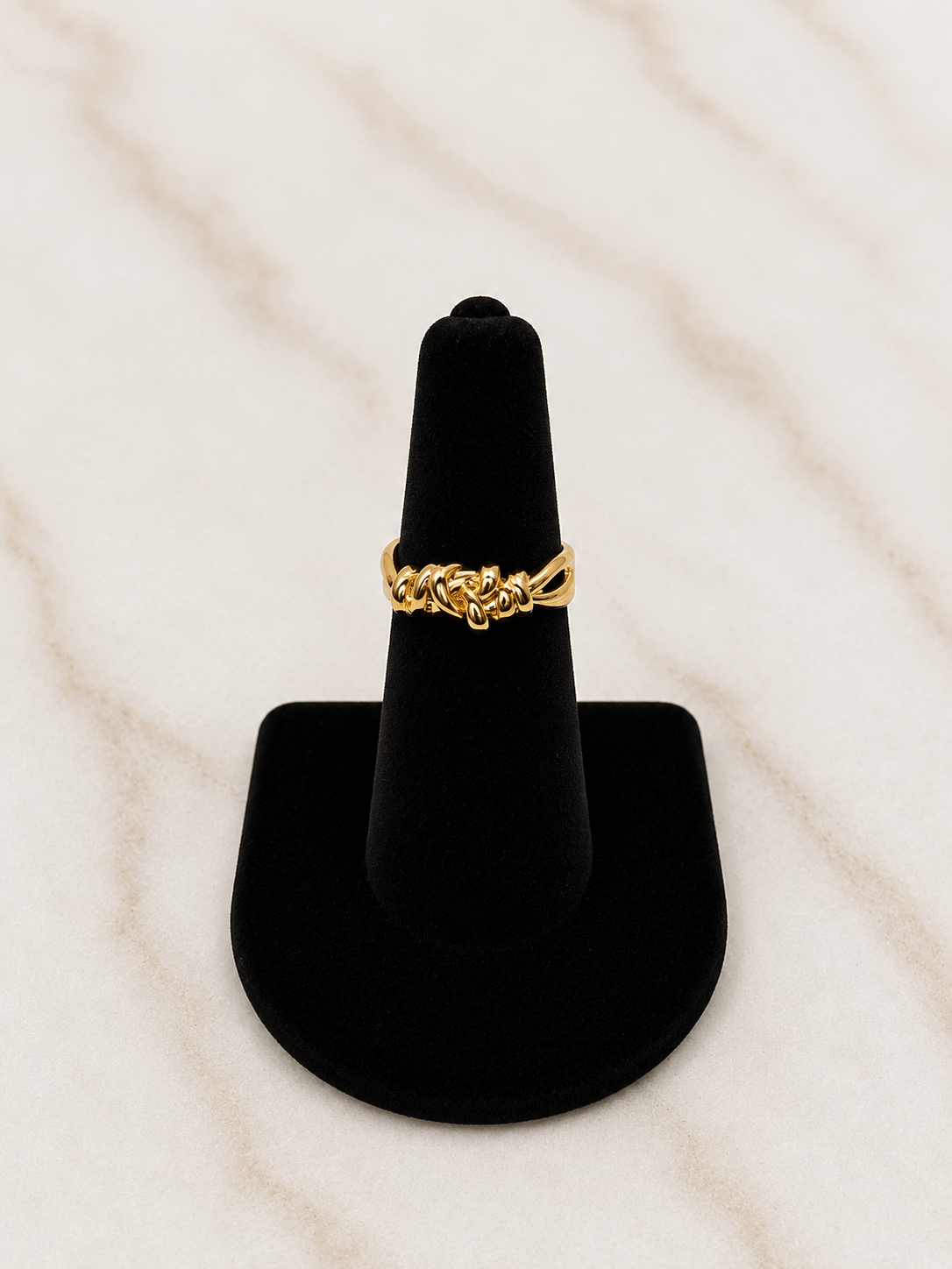 A gold ring with an intricate knot or chain-like design, displayed on a black cone jewelry stand against a light marble background.