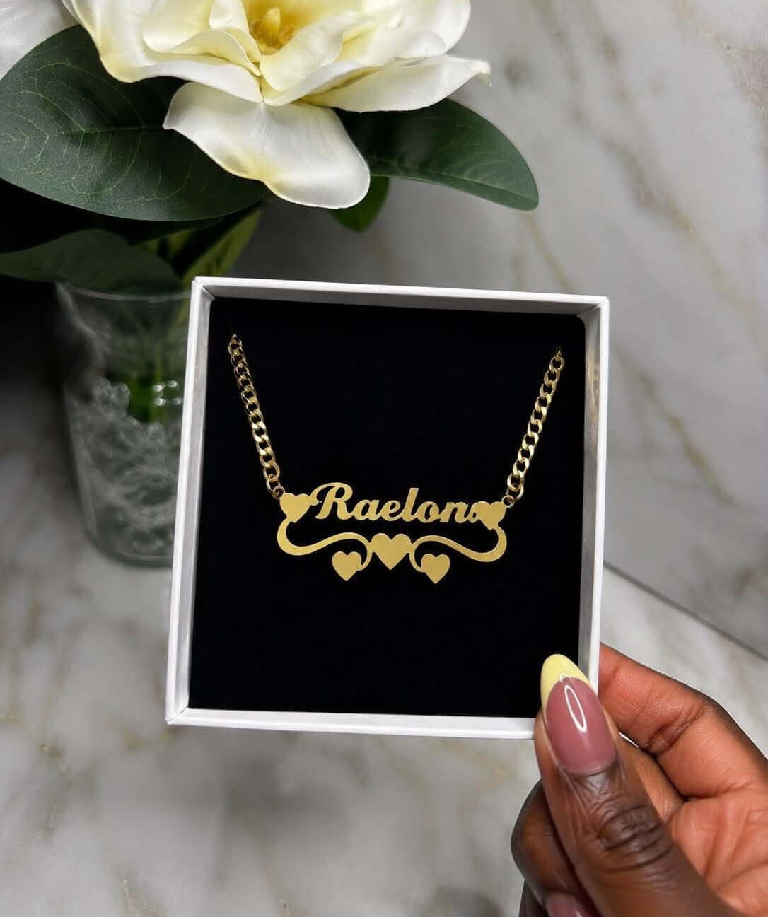 The Reigning Name Necklace displayed in a box with heart details and custom name 'Raelono' in elegant script.