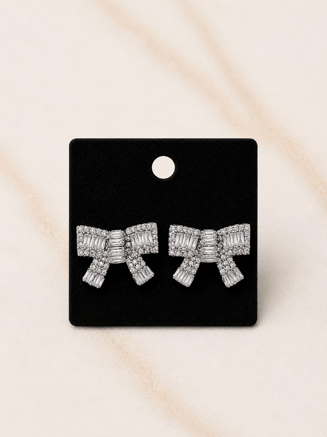 A pair of silver bow-shaped stud earrings paved with clear stones, presented on a black display card against a light beige marble background.