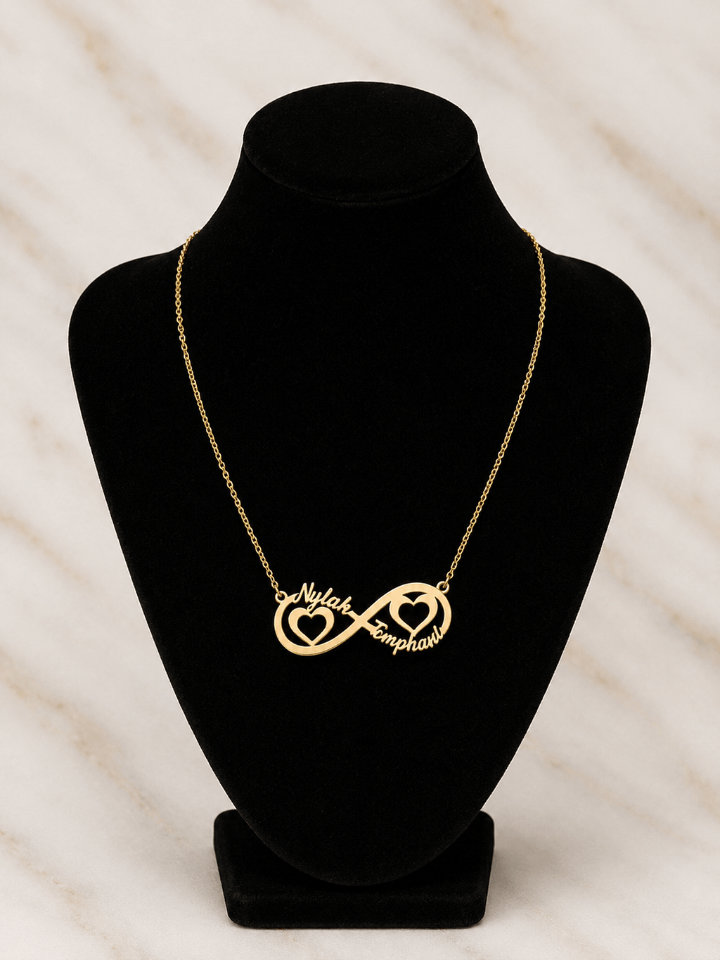 A gold necklace displayed on a black jewelry bust, featuring an infinity symbol with one side shaped like a heart containing the word 'Nail' and the other side shaped like a heart containing the word 'Emphas'.