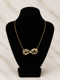 A gold necklace displayed on a black jewelry bust, featuring an infinity symbol with one side shaped like a heart containing the word 'Nail' and the other side shaped like a heart containing the word 'Emphas'.