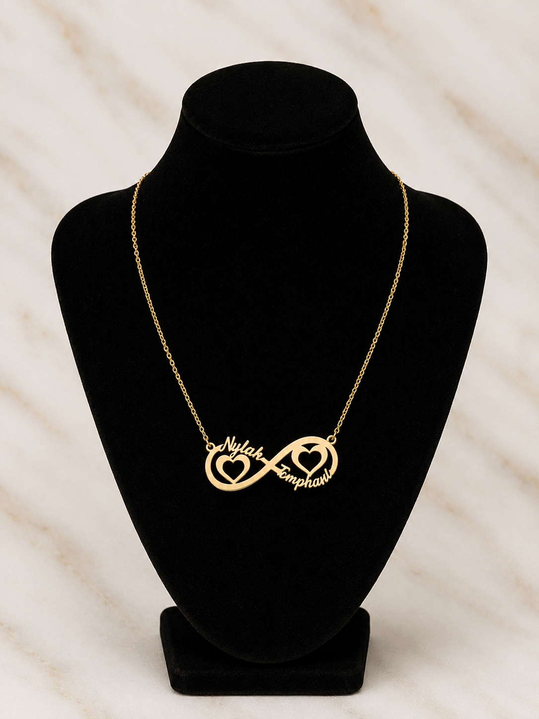 A gold necklace displayed on a black jewelry bust, featuring an infinity symbol with one side shaped like a heart containing the word 'Nail' and the other side shaped like a heart containing the word 'Emphas'.
