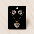 A heart-shaped jewelry set featuring a pendant necklace and matching stud earrings on a black display card. The heart pendants and studs are gold-tone with clear rhinestones around the edges and a central purple and white cartoon character motif.