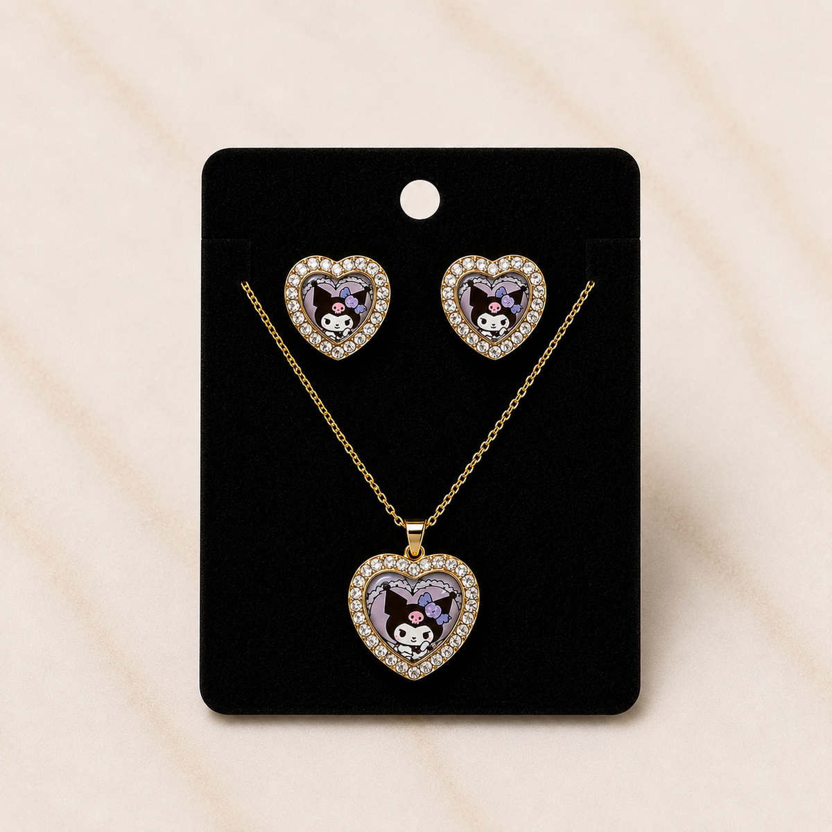 A heart-shaped jewelry set featuring a pendant necklace and matching stud earrings on a black display card. The heart pendants and studs are gold-tone with clear rhinestones around the edges and a central purple and white cartoon character motif.