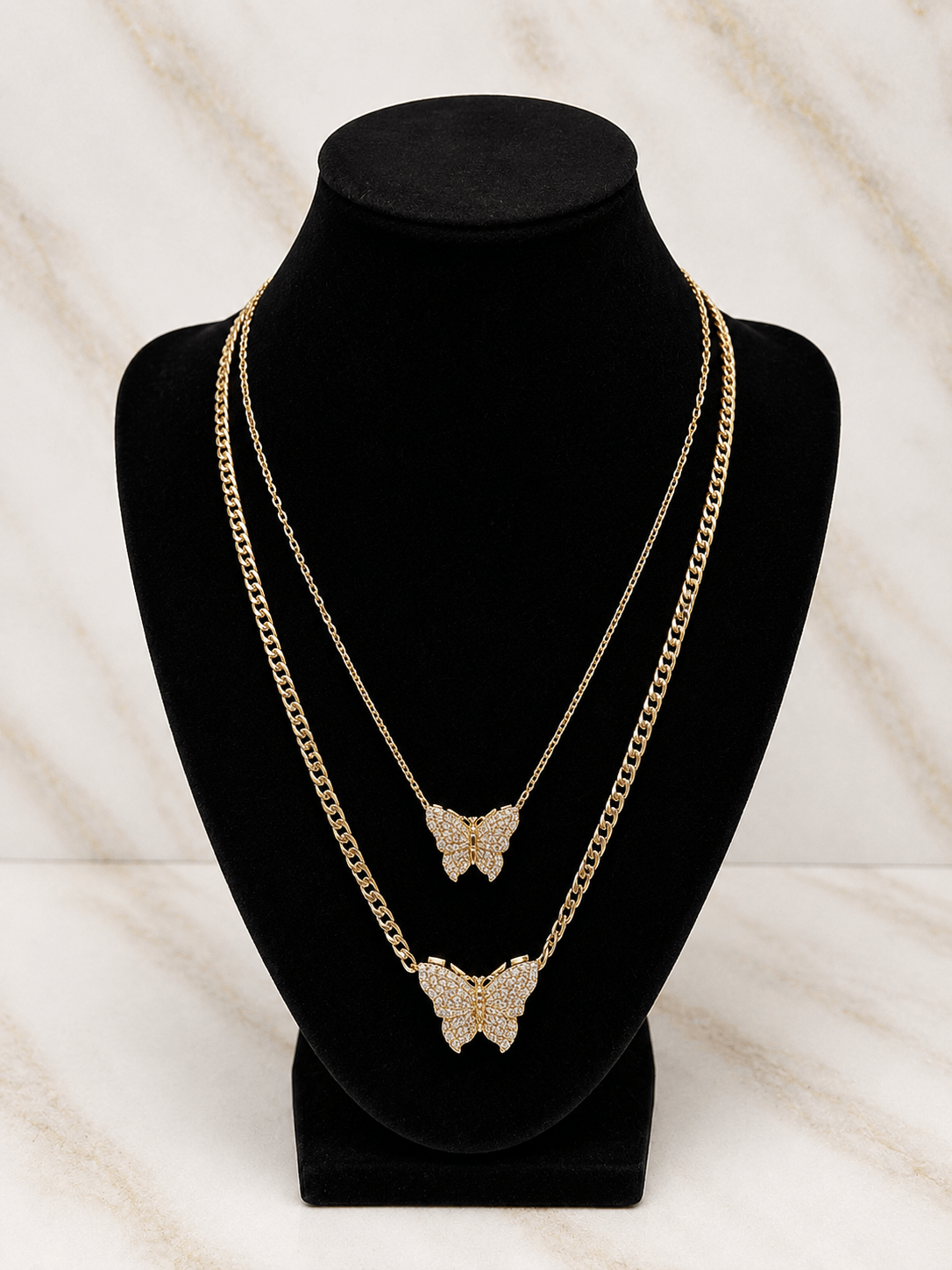 Two layered gold-tone chain necklaces with butterfly pendants, displayed on a black velvet jewelry bust against a light marble-patterned background.
