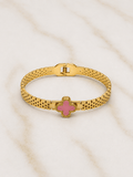 A gold-tone metal bracelet with a pink clover-shaped charm, displayed on a light beige marble surface.