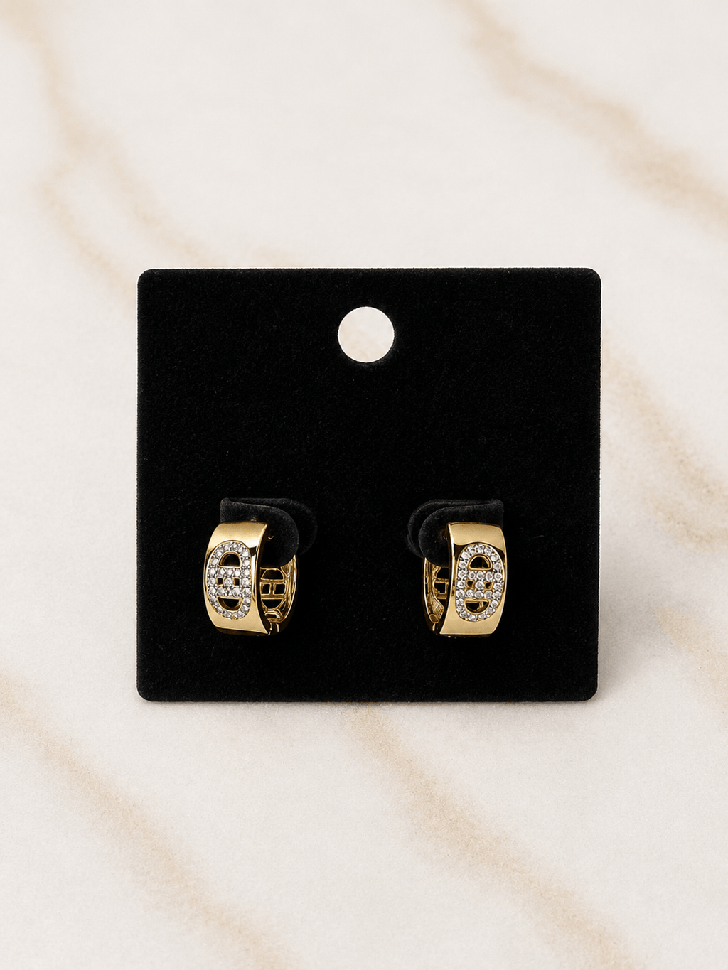 A pair of gold huggie hoop earrings with clear pavé-set stones, displayed on a black velvet card against a light beige marble background.