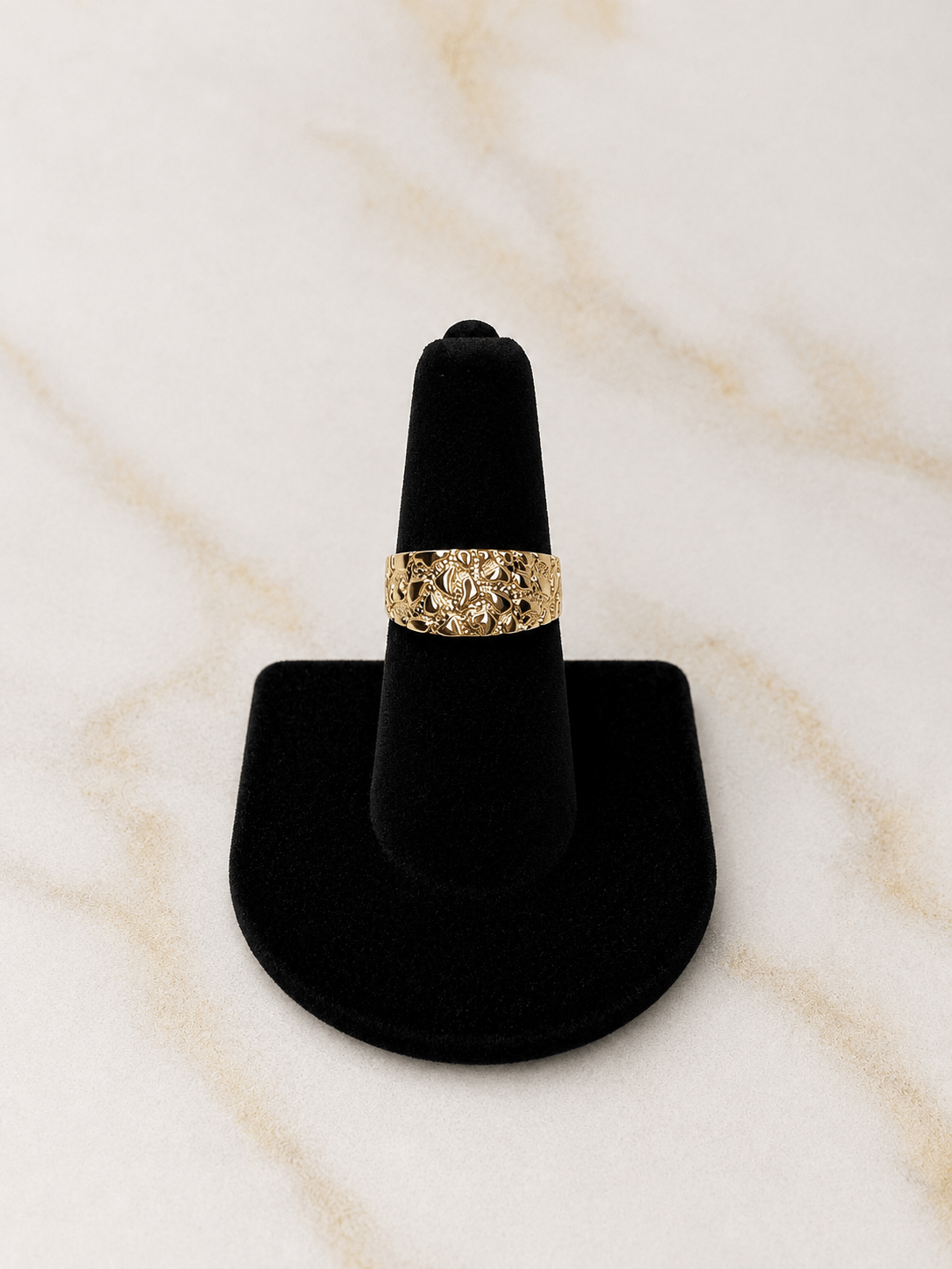 A textured gold ring displayed on a black ring cone stand against a light beige marble background.