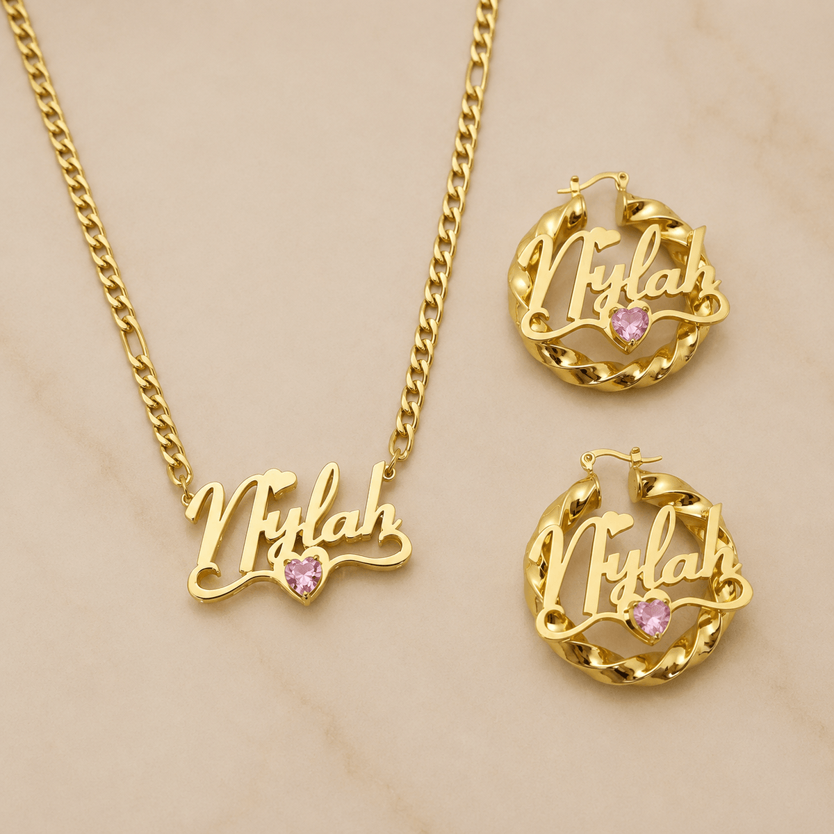 Gold nameplate jewelry set featuring a chain necklace and matching hoop earrings with the name 'Nylah' in cursive script and a small pink heart crystal accent, displayed on a light beige marble background.