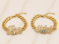 Two gold chain bracelets displayed side by side on a light marble background. The left bracelet is personalized with a nameplate spelling 'Tee' in clear pavé stones and a small clear heart.