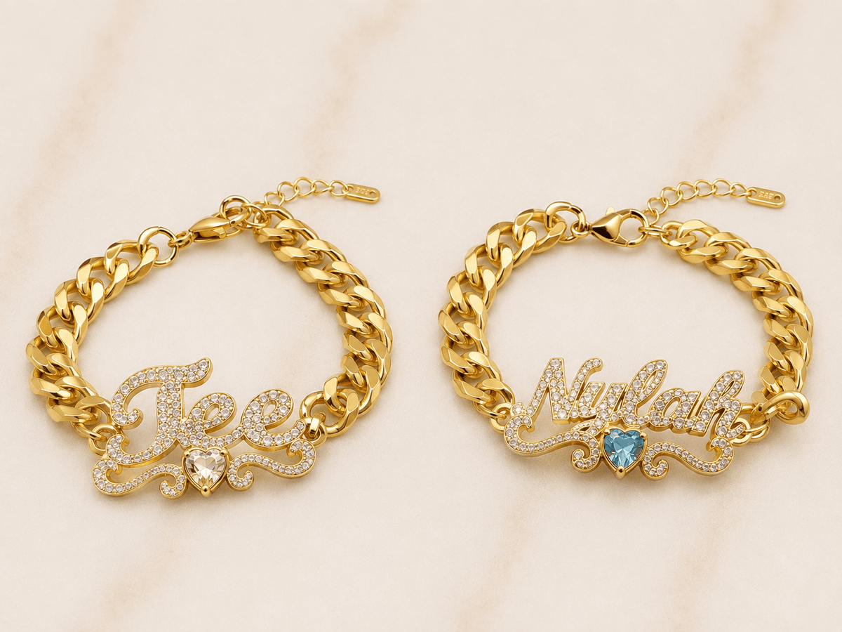 Two gold chain bracelets displayed side by side on a light marble background. The left bracelet is personalized with a nameplate spelling 'Tee' in clear pavé stones and a small clear heart.