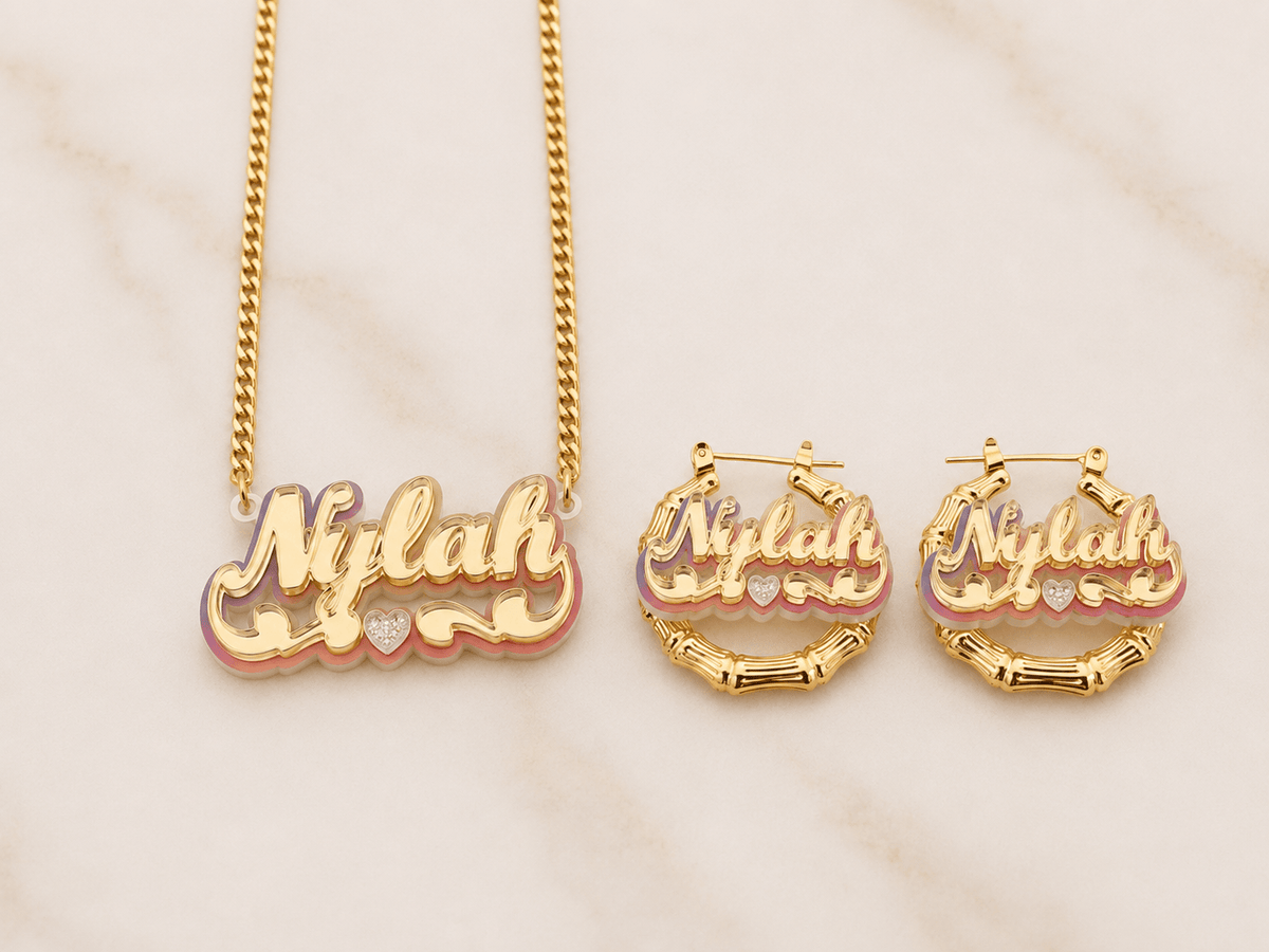 Gold-tone jewelry set featuring a nameplate necklace and matching hoop earrings with the name 'Nylah' in cursive, accented with pink and purple enamel and a small clear rhinestone, displayed on a light marble surface.