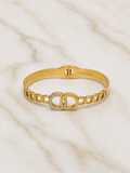 A gold chain bangle bracelet with a central interlocking double-O design, one of which is encrusted with small clear rhinestones, displayed on a light marble surface.