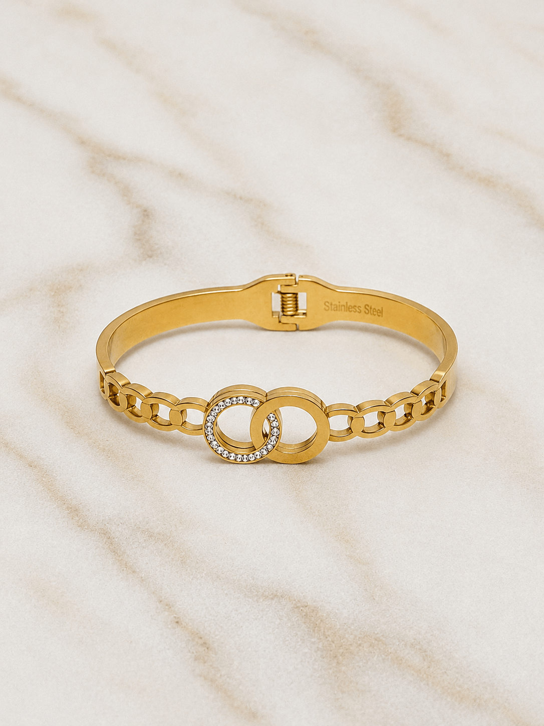 A gold chain bangle bracelet with a central interlocking double-O design, one of which is encrusted with small clear rhinestones, displayed on a light marble surface.