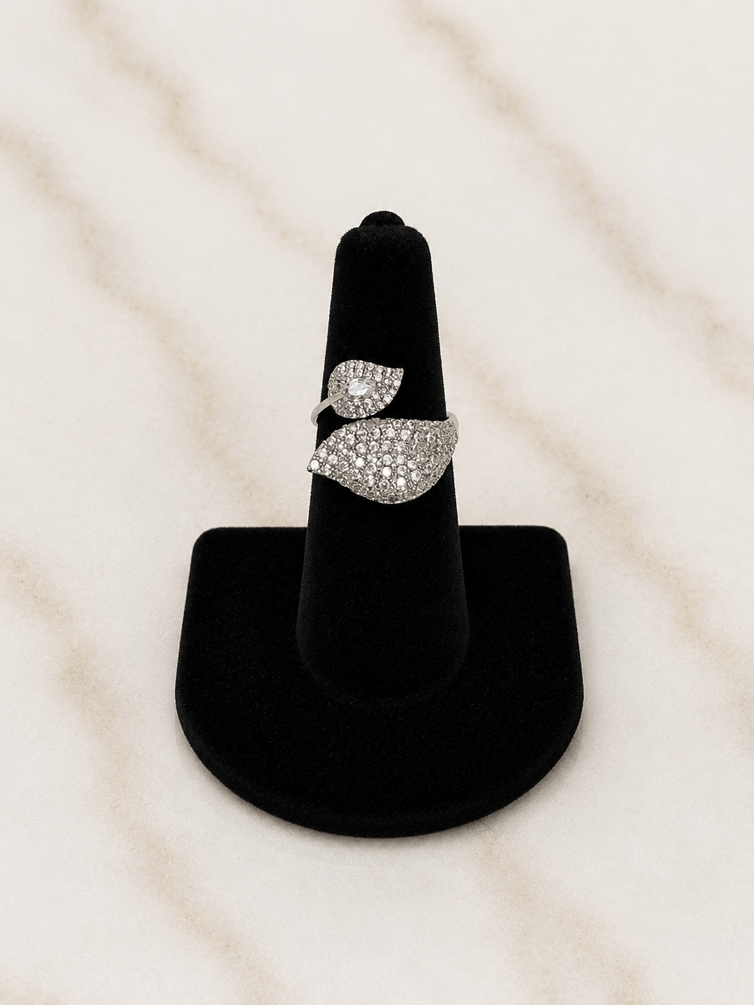 A silver ring with pavé-set clear stones in a leaf-like design, displayed on a black ring stand against a light marble background.
