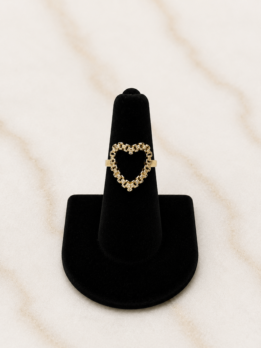 A gold heart-shaped ring displayed on a black velvet ring stand against a light beige marble background.