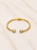 A gold-tone open cuff bangle bracelet with small clear baguette gemstones at each end, displayed on a light beige marble surface.