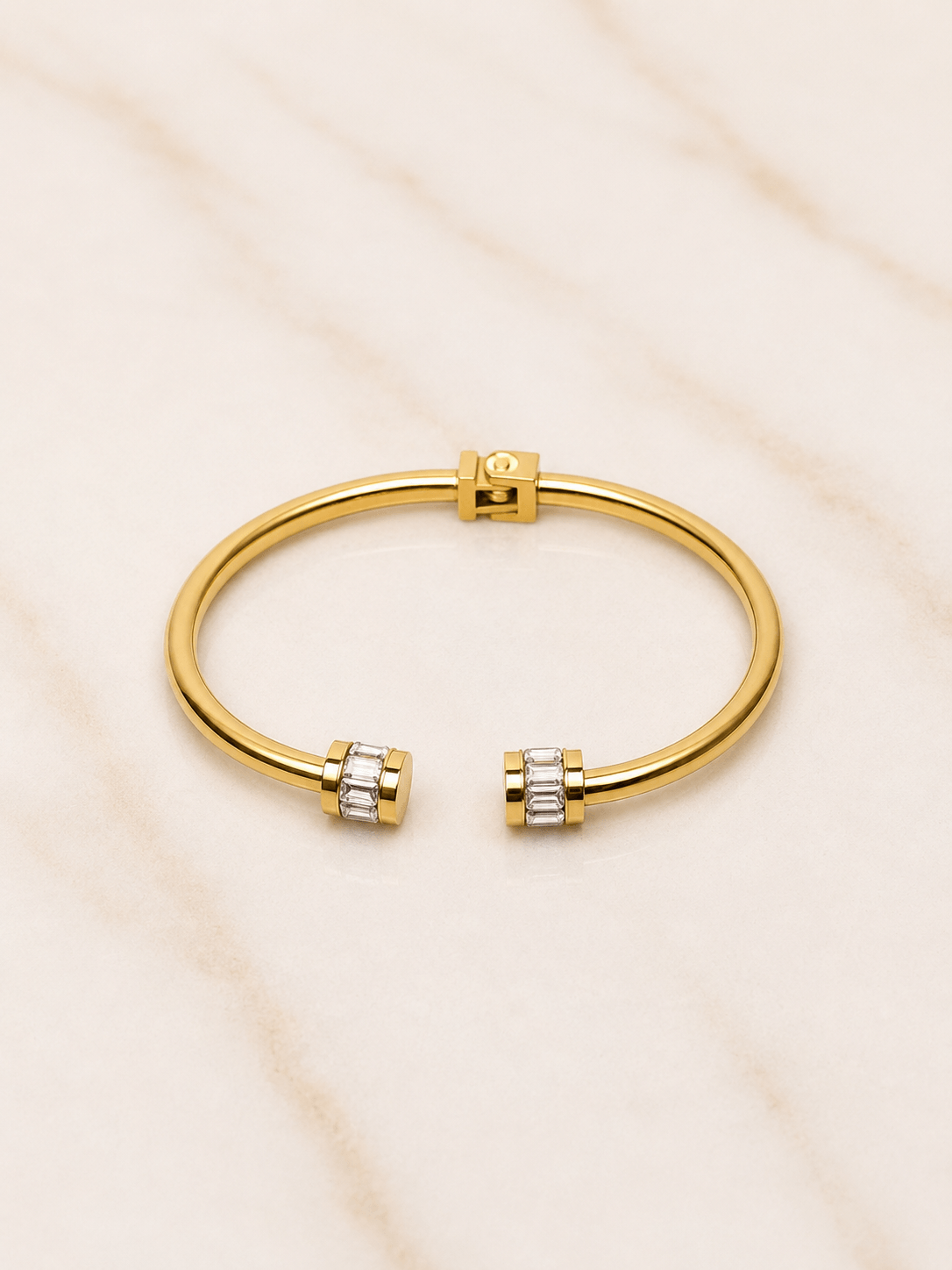 A gold-tone open cuff bangle bracelet with small clear baguette gemstones at each end, displayed on a light beige marble surface.