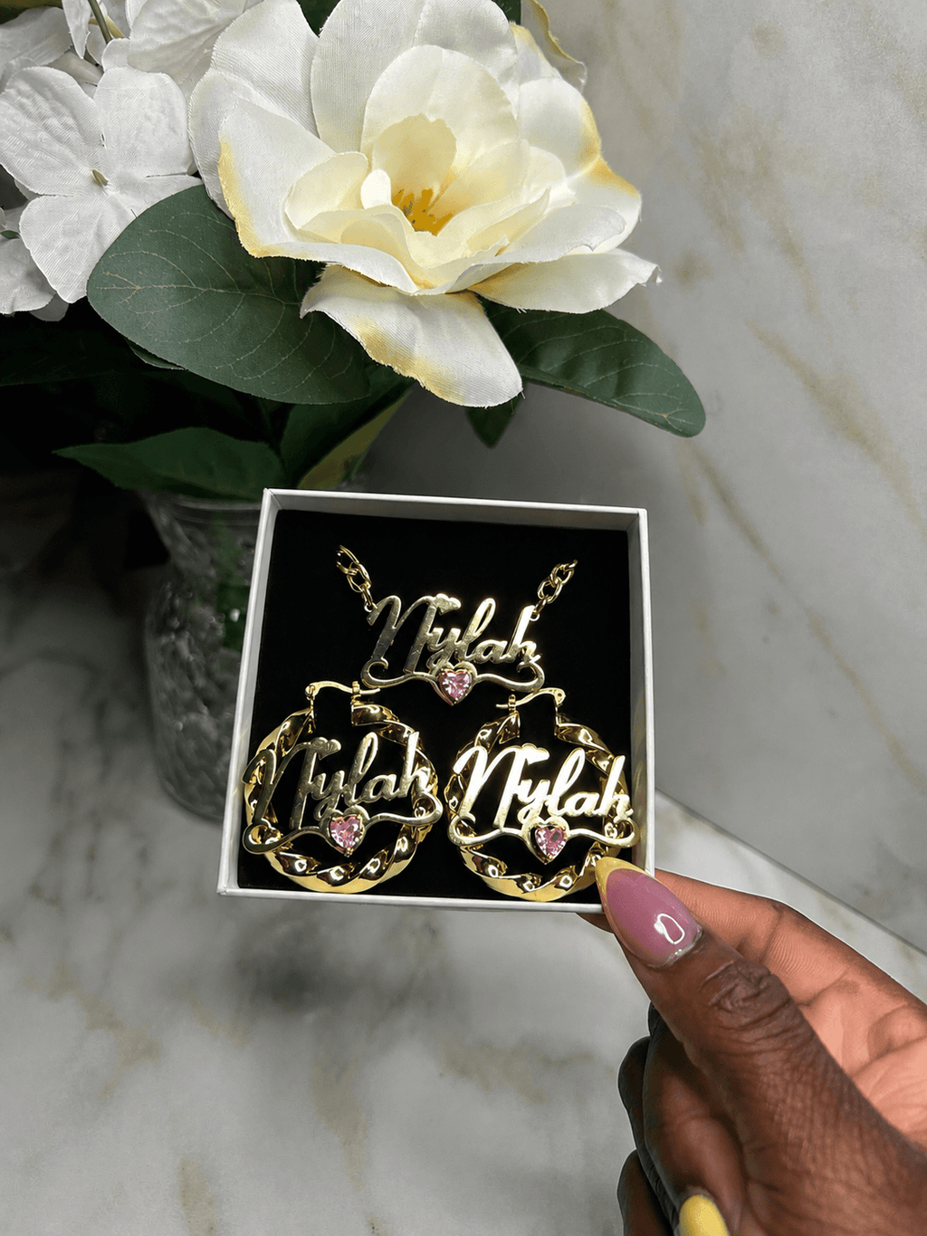 A hand holds an open jewelry box containing two gold-tone necklaces with the name 'Niyah' in cursive and a matching larger pendant, each with a small pink heart detail. White flowers and green leaves are in the background against a light marble surface.