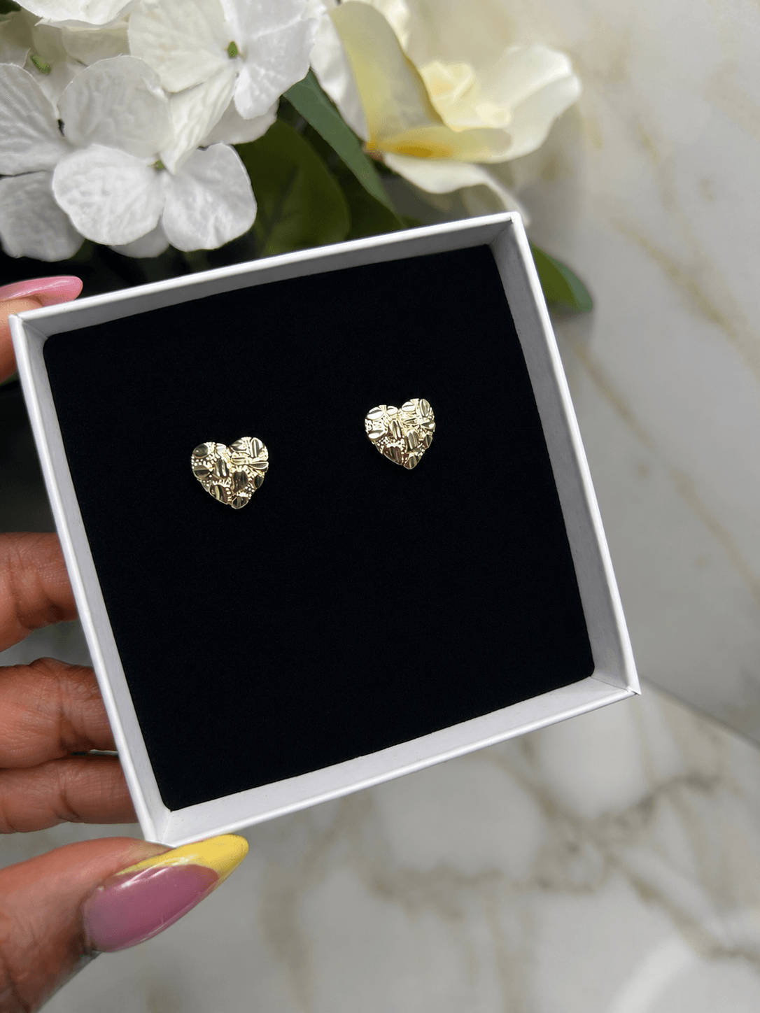 A pair of textured gold heart stud earrings displayed on a black velvet insert inside an open white gift box, held by a hand with pink and yellow nail polish against a marble surface and white flowers.