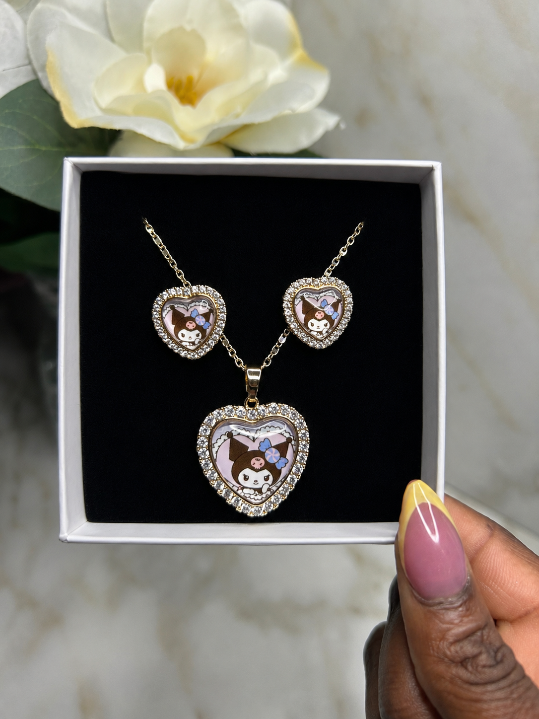 A heart-shaped pendant necklace and matching heart-shaped earrings displayed in an open white gift box with a black insert. The jewelry has a gold-tone outline, clear rhinestone borders, and cartoon character enamel details.