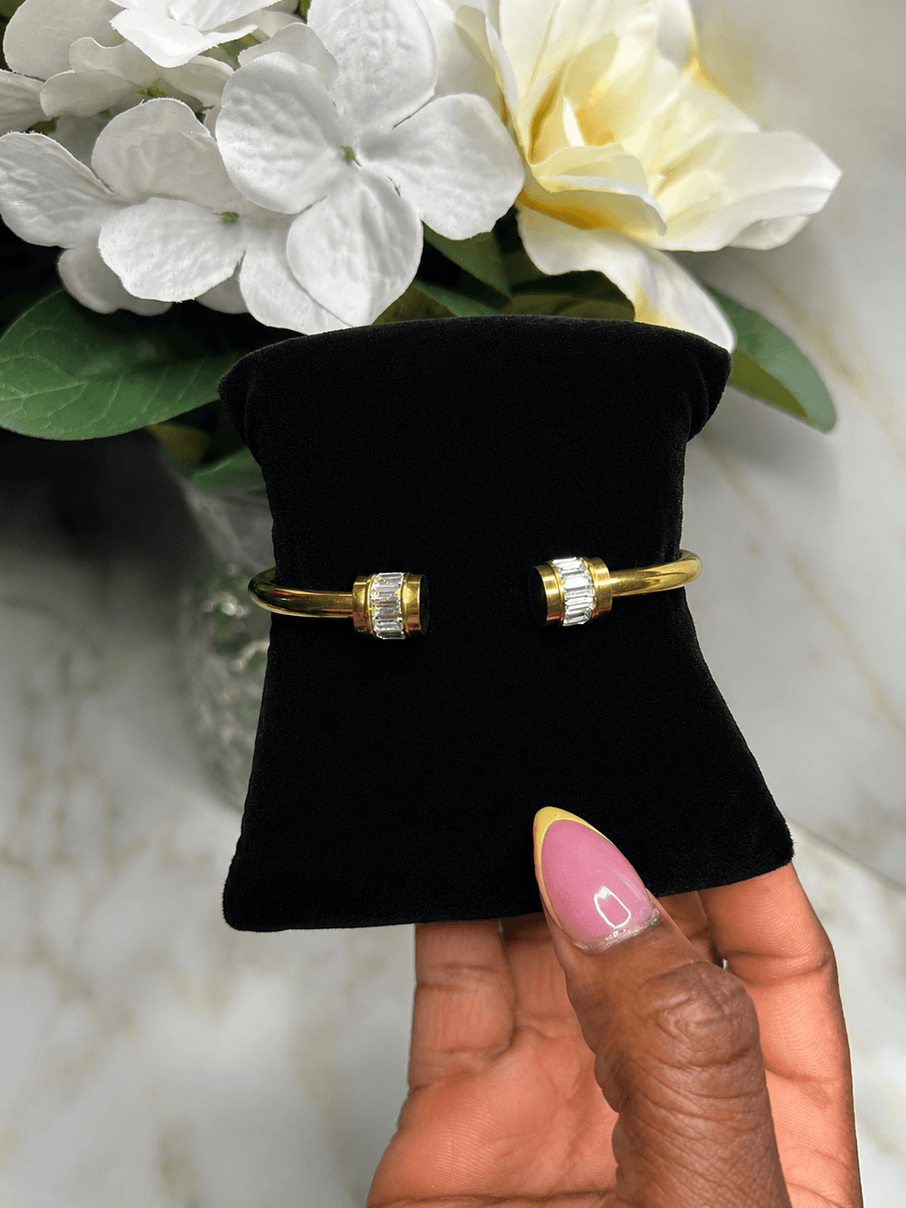 A pair of gold-toned cuff bracelets with clear rectangular gemstones, displayed on a black velvet jewelry stand held by a hand with pink and yellow nail polish, with white flowers in the background.