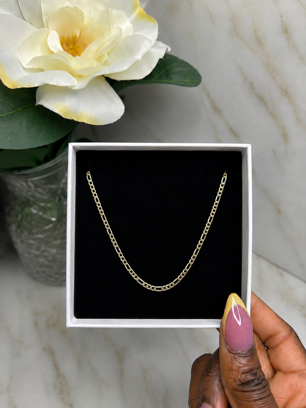 A gold chain necklace displayed inside a white jewelry box with a black insert, held by a hand with pink and yellow nail art, with a white flower in a vase in the background.