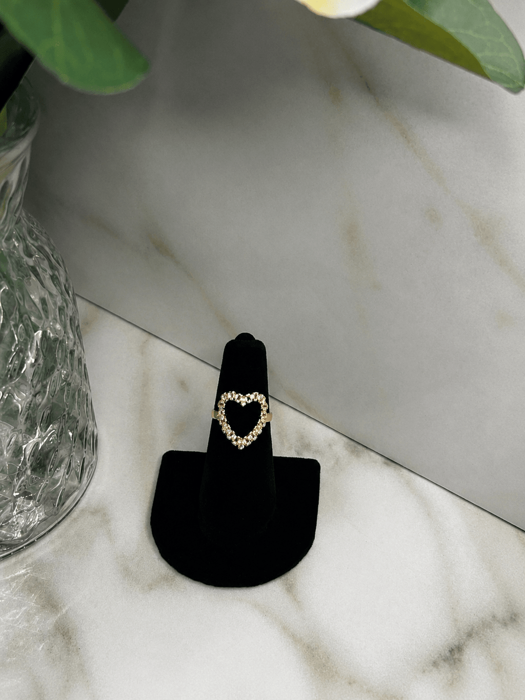 A gold heart-shaped ring displayed on a black ring stand, set on a white marble surface next to a glass vase.