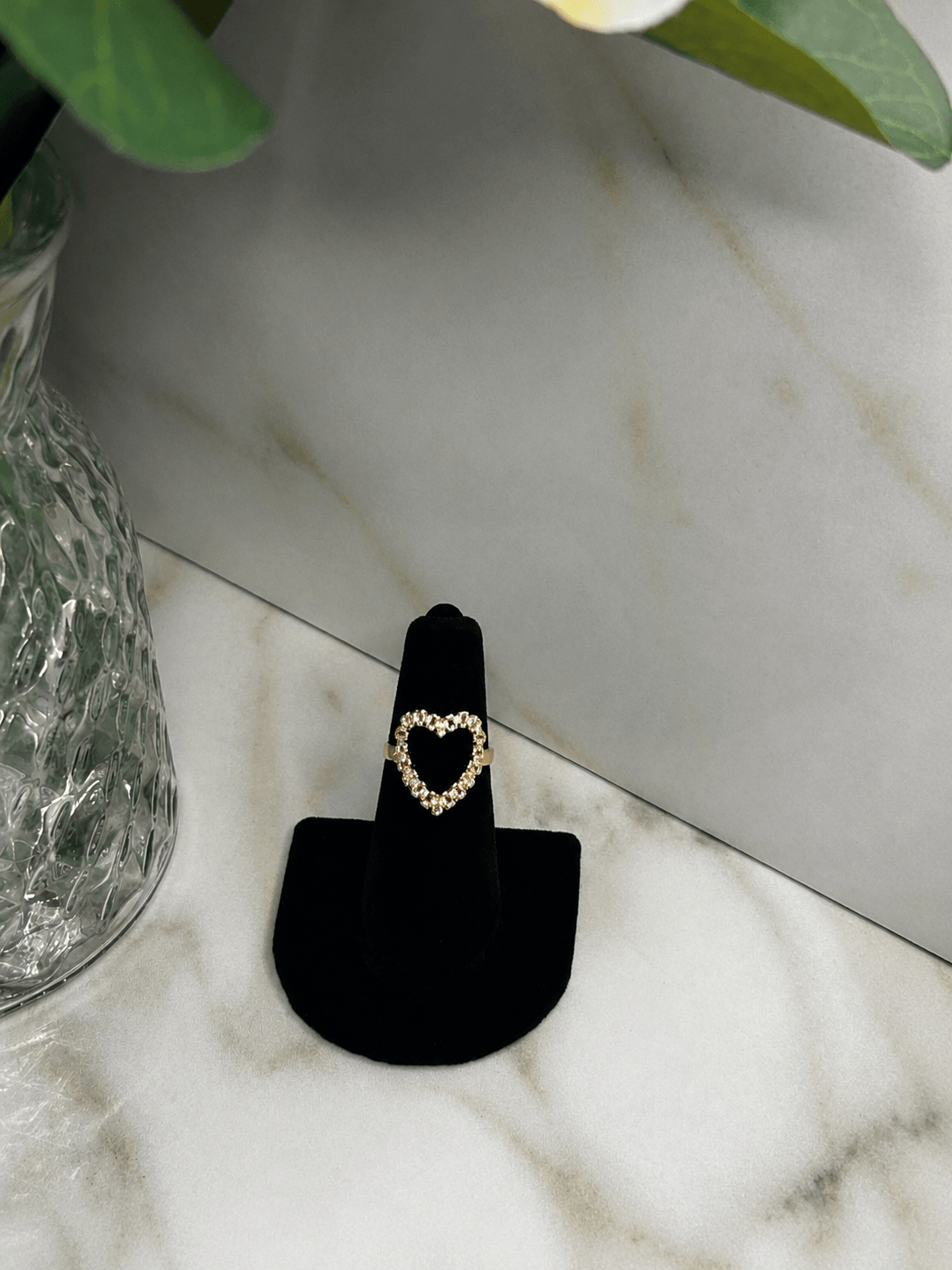 A gold heart-shaped ring displayed on a black ring stand, set on a white marble surface next to a glass vase.