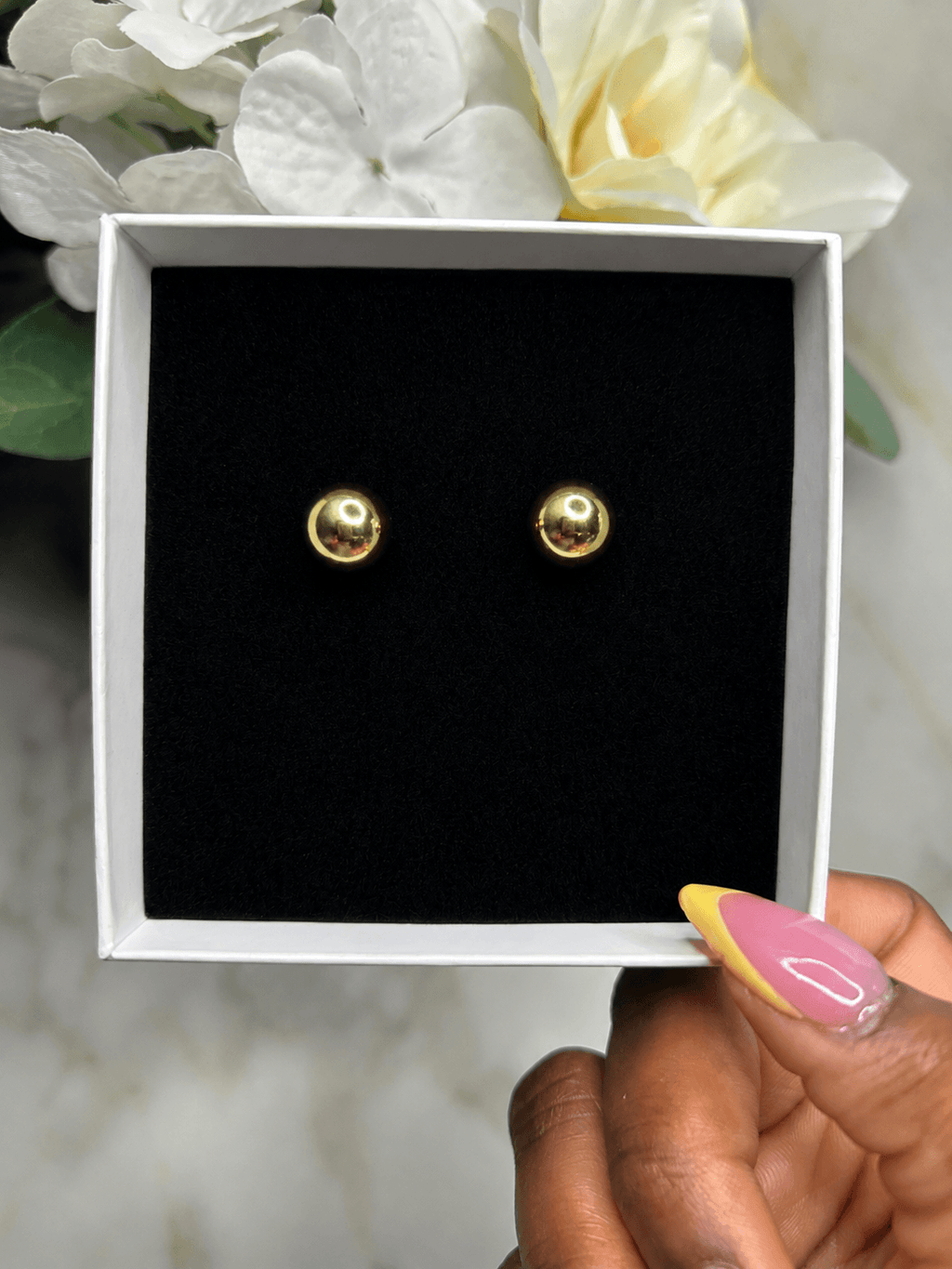 A pair of small gold stud earrings displayed inside an open white jewelry box with a black velvet insert, held by a hand with pink and yellow manicured nails, with white flowers in the background.