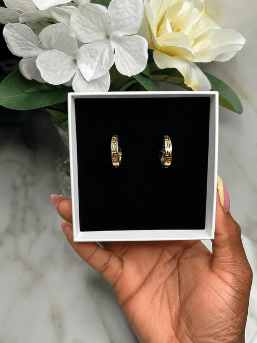 A hand holds a white jewelry box with a black velvet insert displaying a pair of gold hoop earrings with geometric cutout details. White and yellow flowers are in the background on a marble surface.