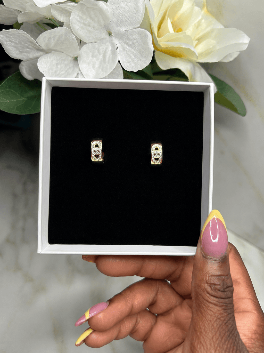 A pair of gold-toned rectangular stud earrings with clear stones, displayed on a black insert inside an open white gift box, held by a hand with pink and yellow manicured nails.