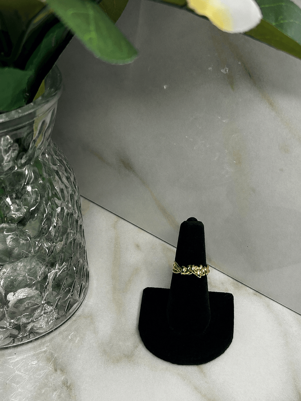 A gold ring displayed on a black ring stand, set on a light marble surface next to a glass vase with green leaves.