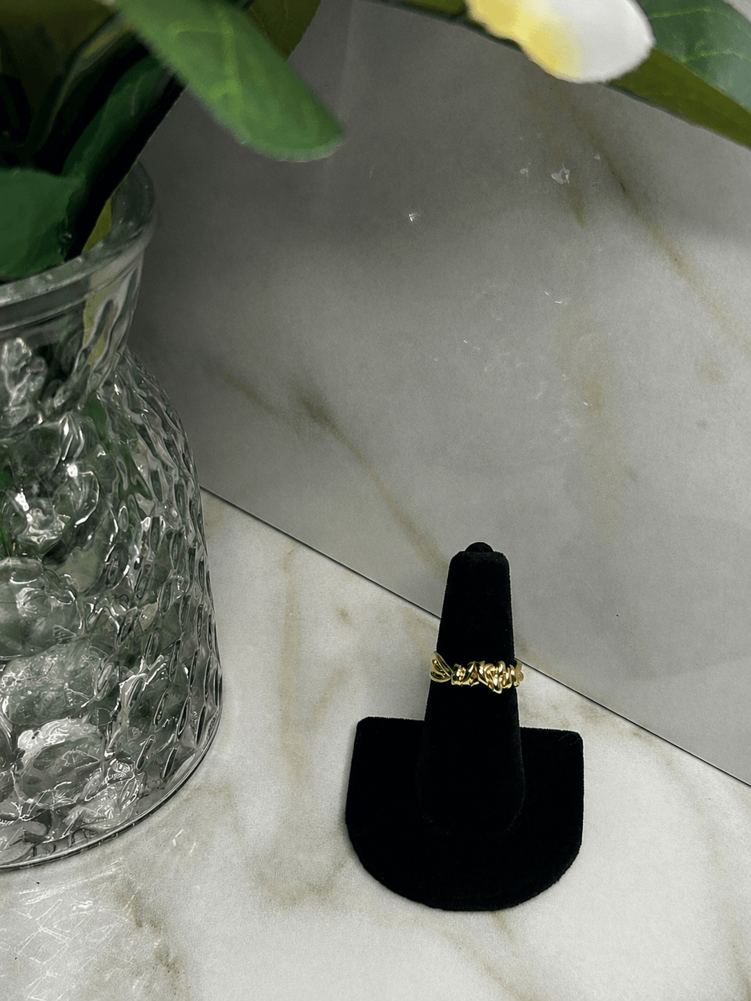 A gold ring displayed on a black ring stand, set on a light marble surface next to a glass vase with green leaves.