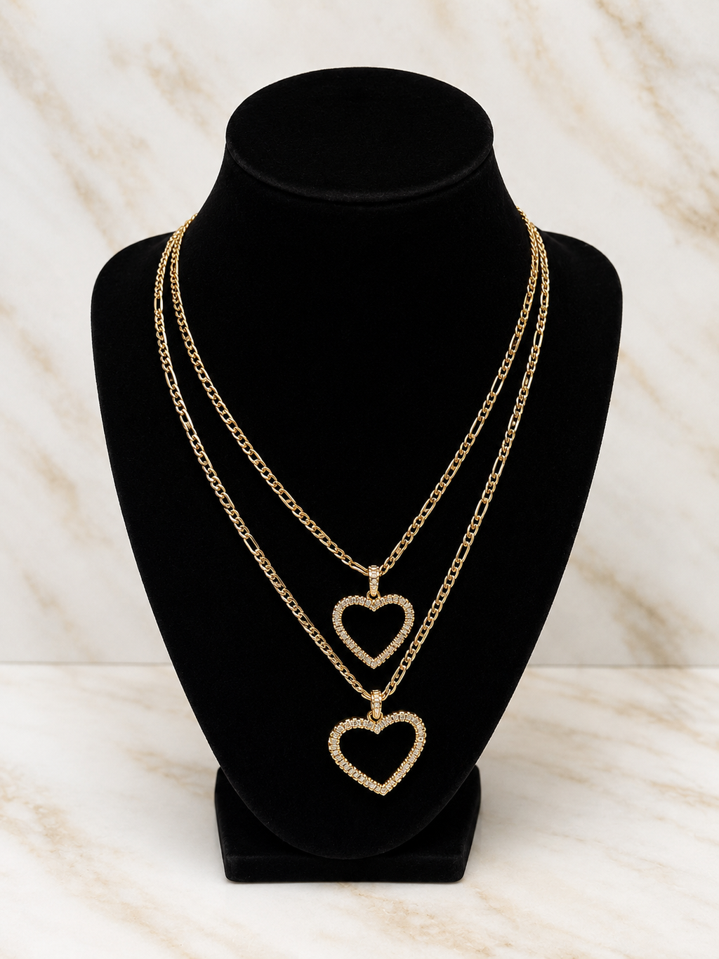 Two gold-tone layered necklaces displayed on a black velvet jewelry bust against a light marble-patterned background. The necklaces feature open heart pendants, with the top layer in a chain link style and the bottom layer in a figure-eight style.