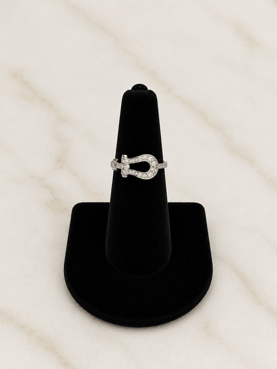 A silver ring with a horseshoe-shaped pavé setting, displayed on a black cone stand against a light marble background.