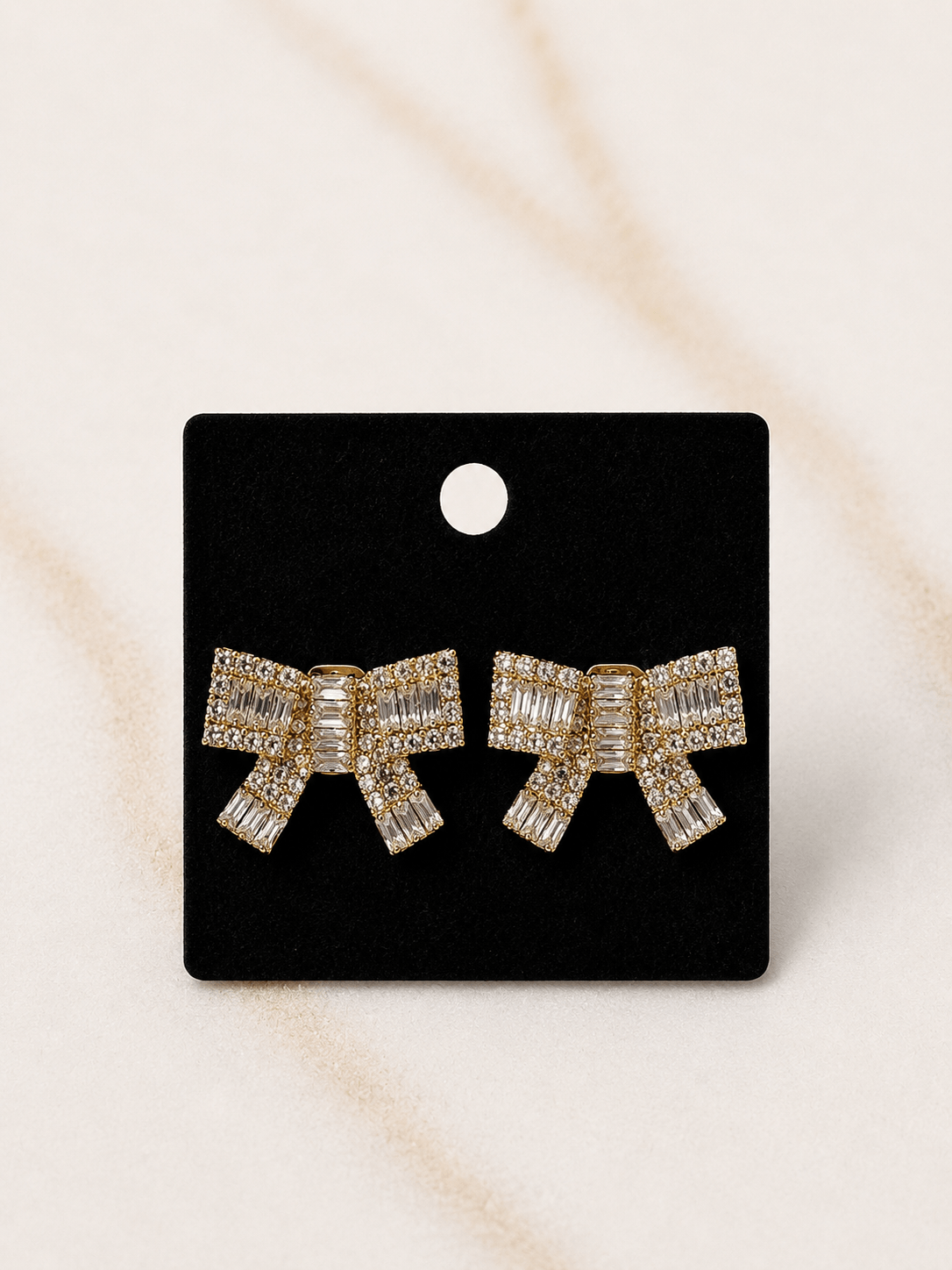 A pair of gold bow-shaped stud earrings encrusted with clear baguette and round crystals, presented on a black display card against a light beige marble background.
