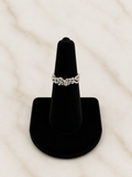 A silver-toned ring with an intricate floral or vine pattern, displayed on a black velvet ring cone stand against a light marble background.