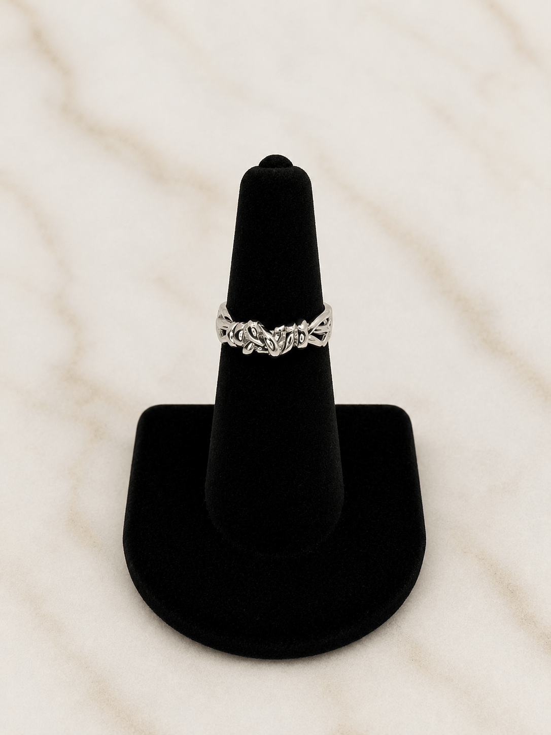 A silver-toned ring with an intricate floral or vine pattern, displayed on a black velvet ring cone stand against a light marble background.