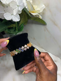 A close-up of a hand holding a black velvet jewelry display pad with a colorful floral bracelet on it. The bracelet features multiple small flower motifs in green, blue, pink, orange, and yellow on a gold-toned band.