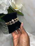 A gold-toned chain bracelet with floral and crystal details displayed on a black velvet jewelry stand, held by a hand against a marble surface with white flowers in the background.