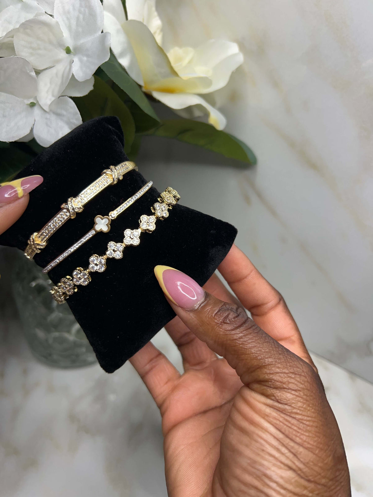 A hand with pink and yellow manicured nails holds a black velvet jewelry display stand featuring four gold-toned bracelets, set against a marble background with white flowers.