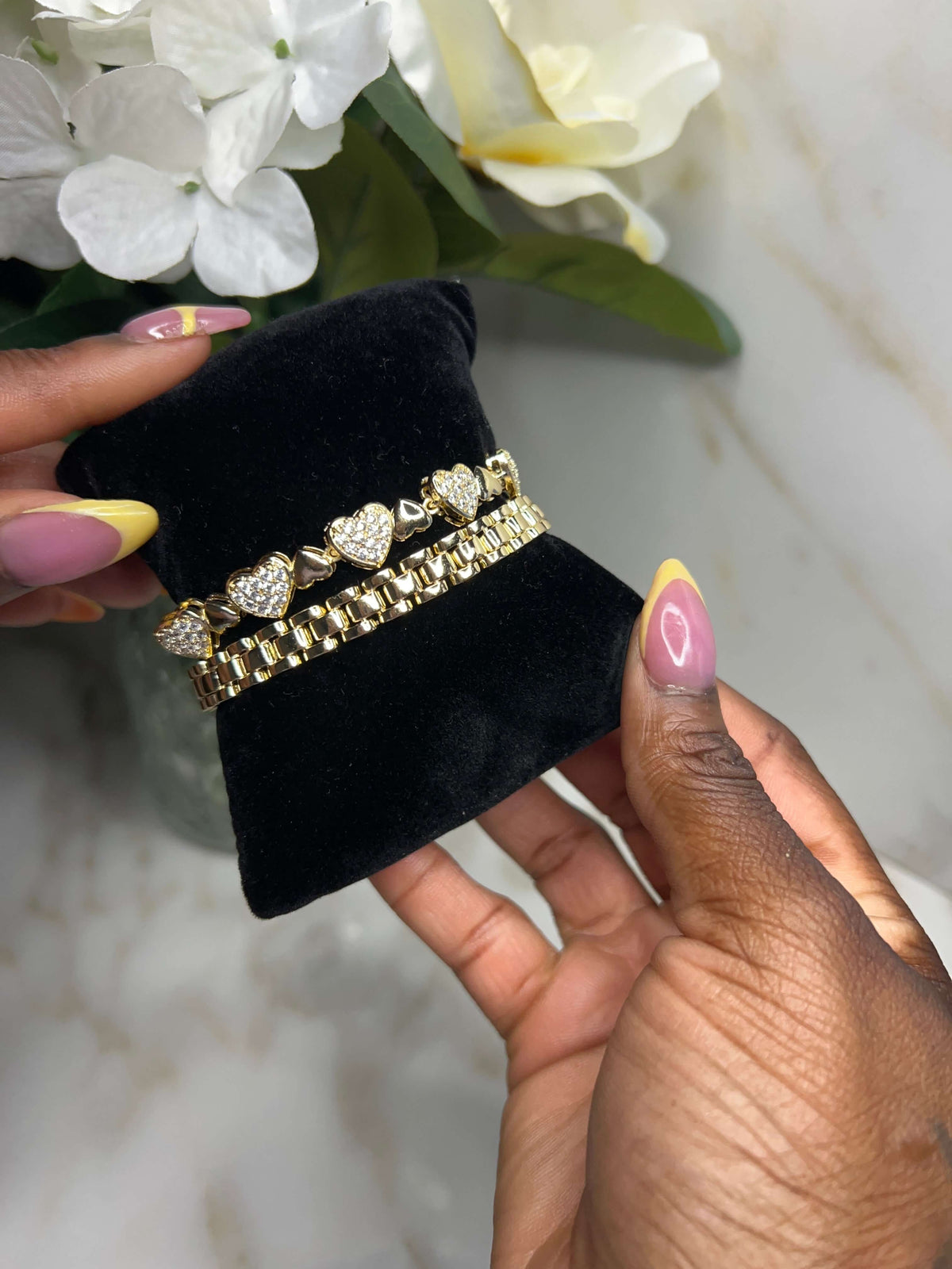 A close-up of a person holding a black velvet jewelry display cushion with two gold bracelets featuring heart-shaped crystal details, set against a marble background with white flowers.